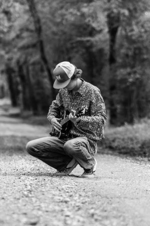 Black and white photo of someone sitting on gravel path playing acoustic guitar in a floral print shirt.