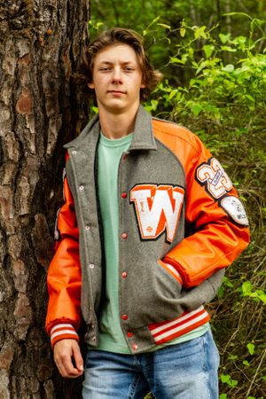 Person wearing orange and grey varsity jacket with jeans leaning against tree trunk in outdoor setting.