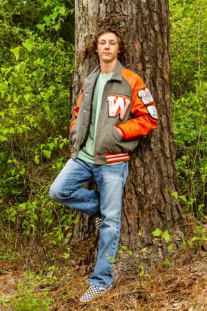 Casual portrait against a tree trunk wearing varsity jacket and checkered shoes.