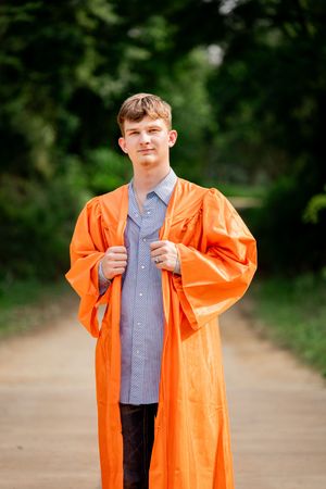 Young man in orange graduation gown standing on a tree-lined path, smiling confidently at the camera.