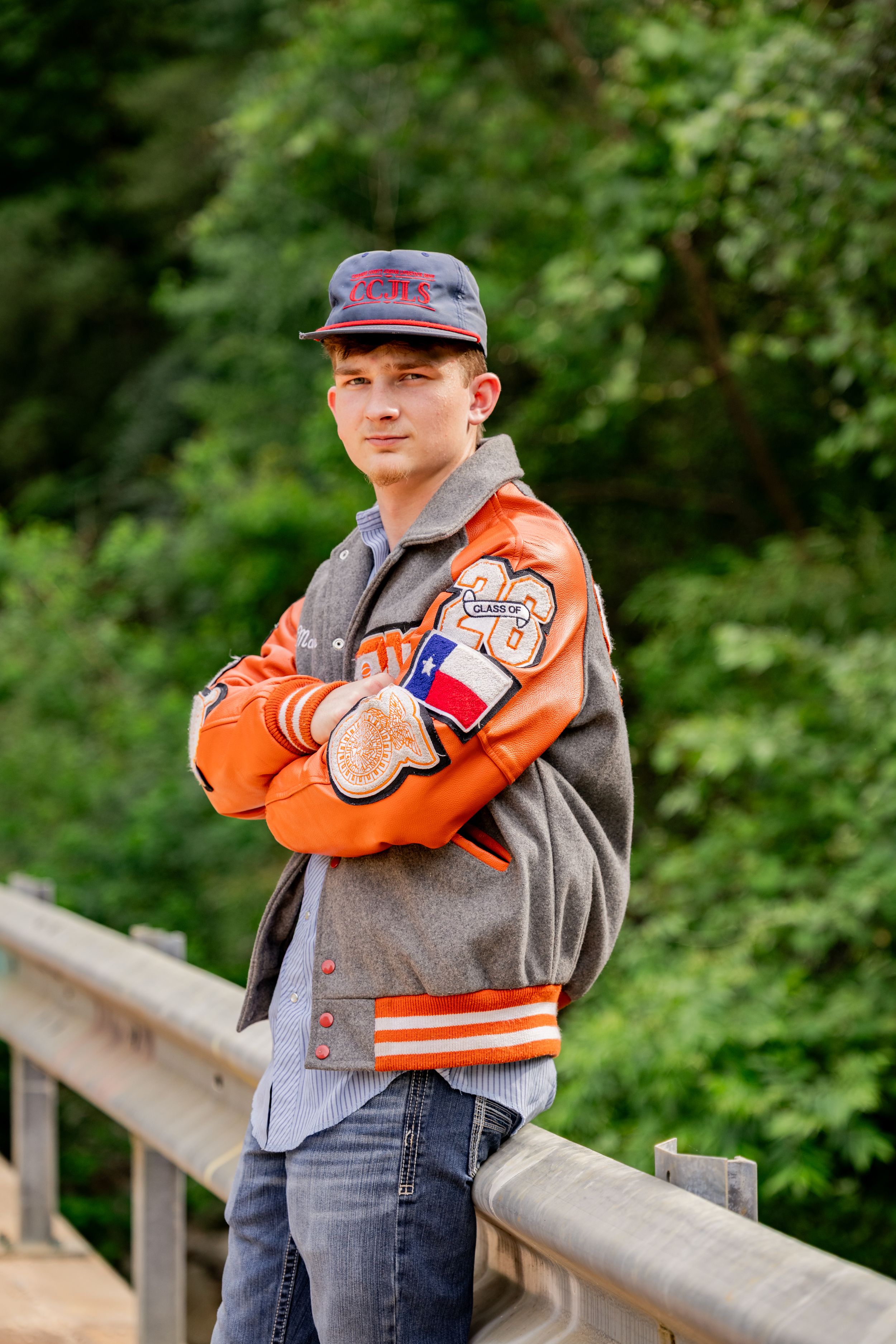 Teen boy in orange varsity jacket and cap poses arms crossed on wooden bridge surrounded by green trees.