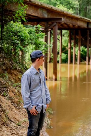 Young man in blue shirt and cap standing by a muddy river beneath a rustic wooden bridge in a forest.