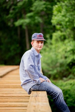 Teen boy in blue striped shirt and cap sitting on a wooden bridge railing surrounded by lush green trees.