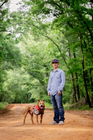A young man in a blue shirt and cap stands with a brown pit bull on a red dirt forest trail.