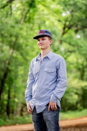 Young man in blue striped shirt and baseball cap smiling outdoors in a lush green forest setting.