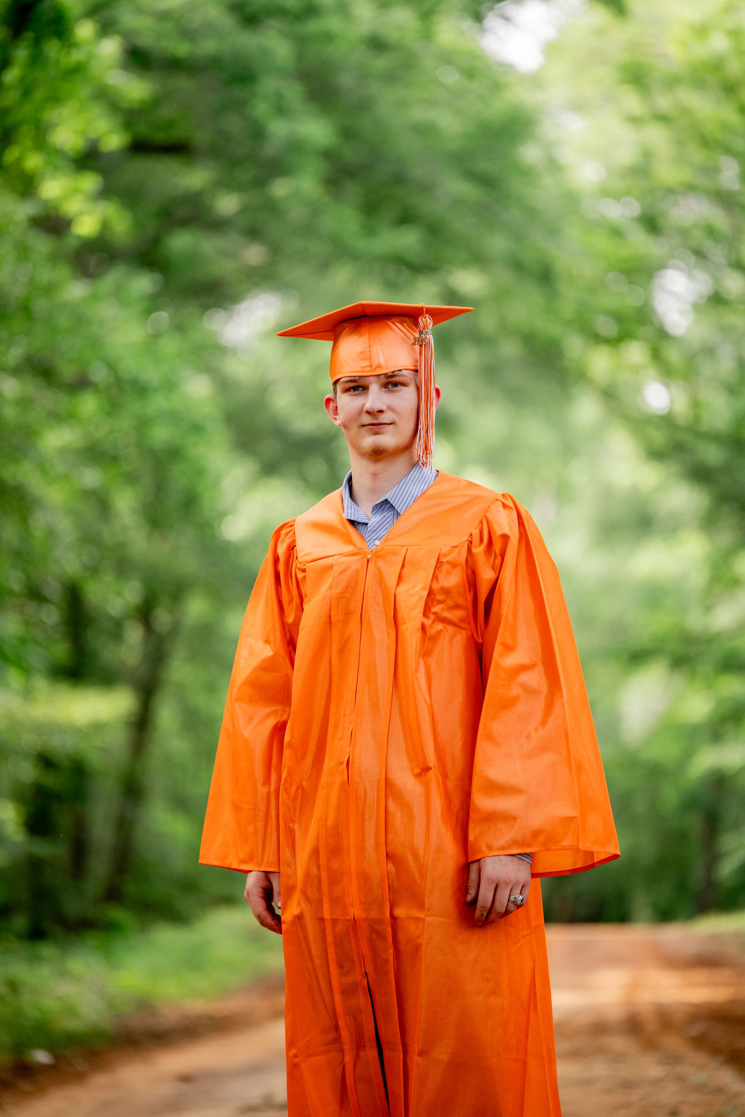Young man in orange graduation cap and gown standing on a dirt path surrounded by green trees.