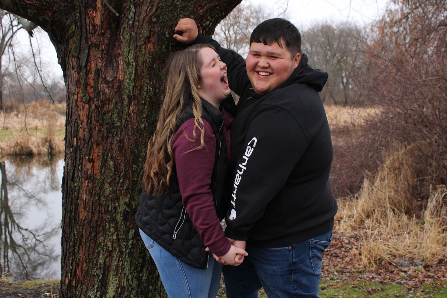 A young couple laughing and holding hands by a tree near a pond in a park during autumn.