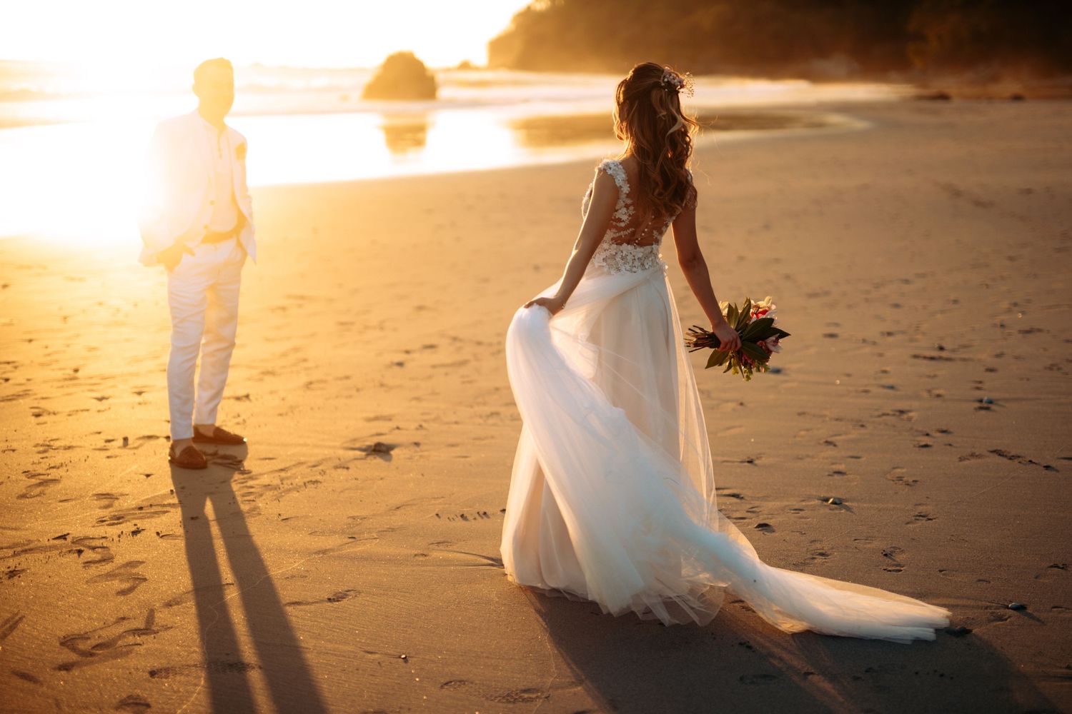 A romantic beach wedding photo at sunset with silhouettes and flowing white dress.