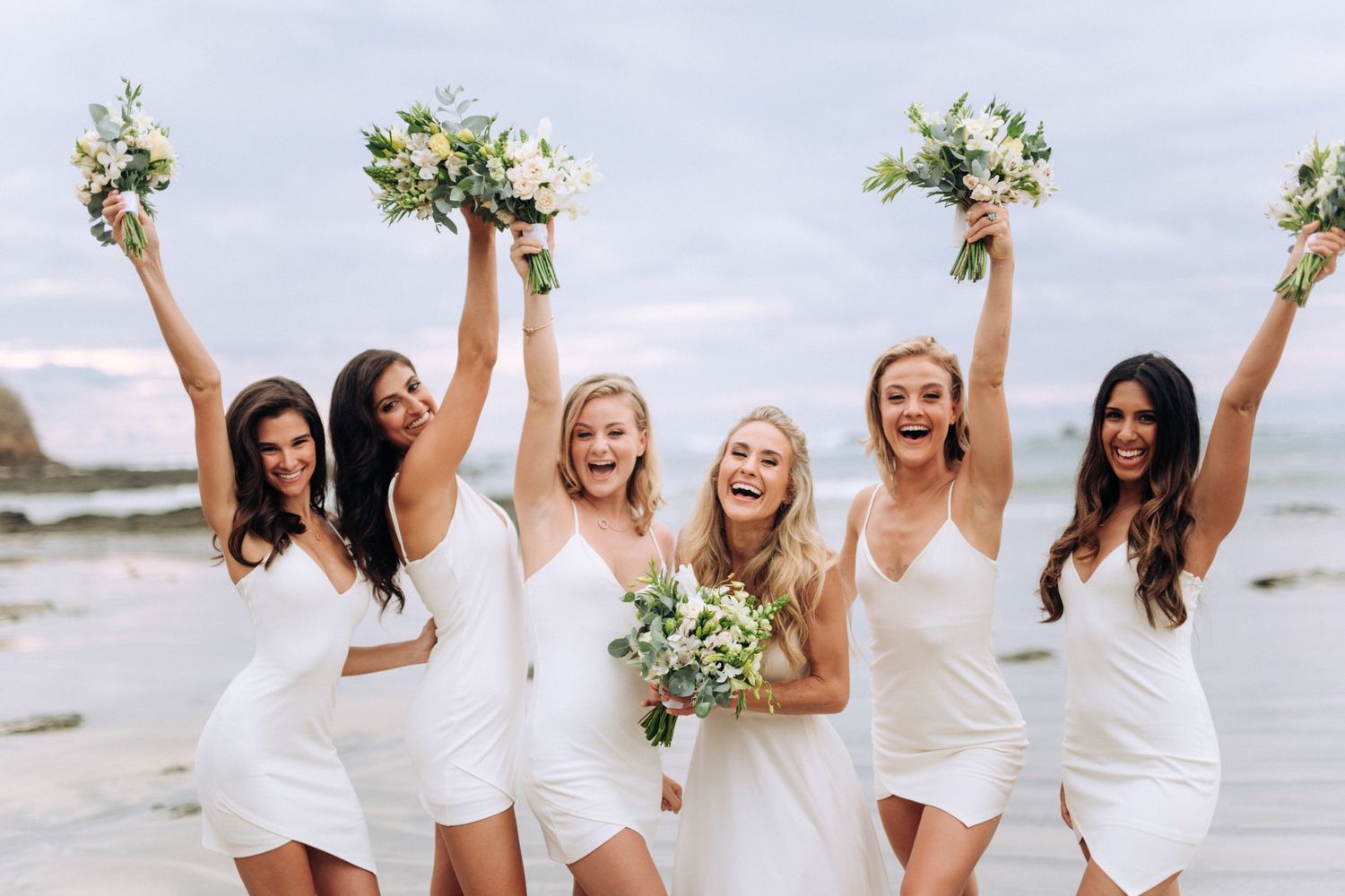 A joyful bridal party in white dresses celebrating on a beach while holding floral bouquets in the air.