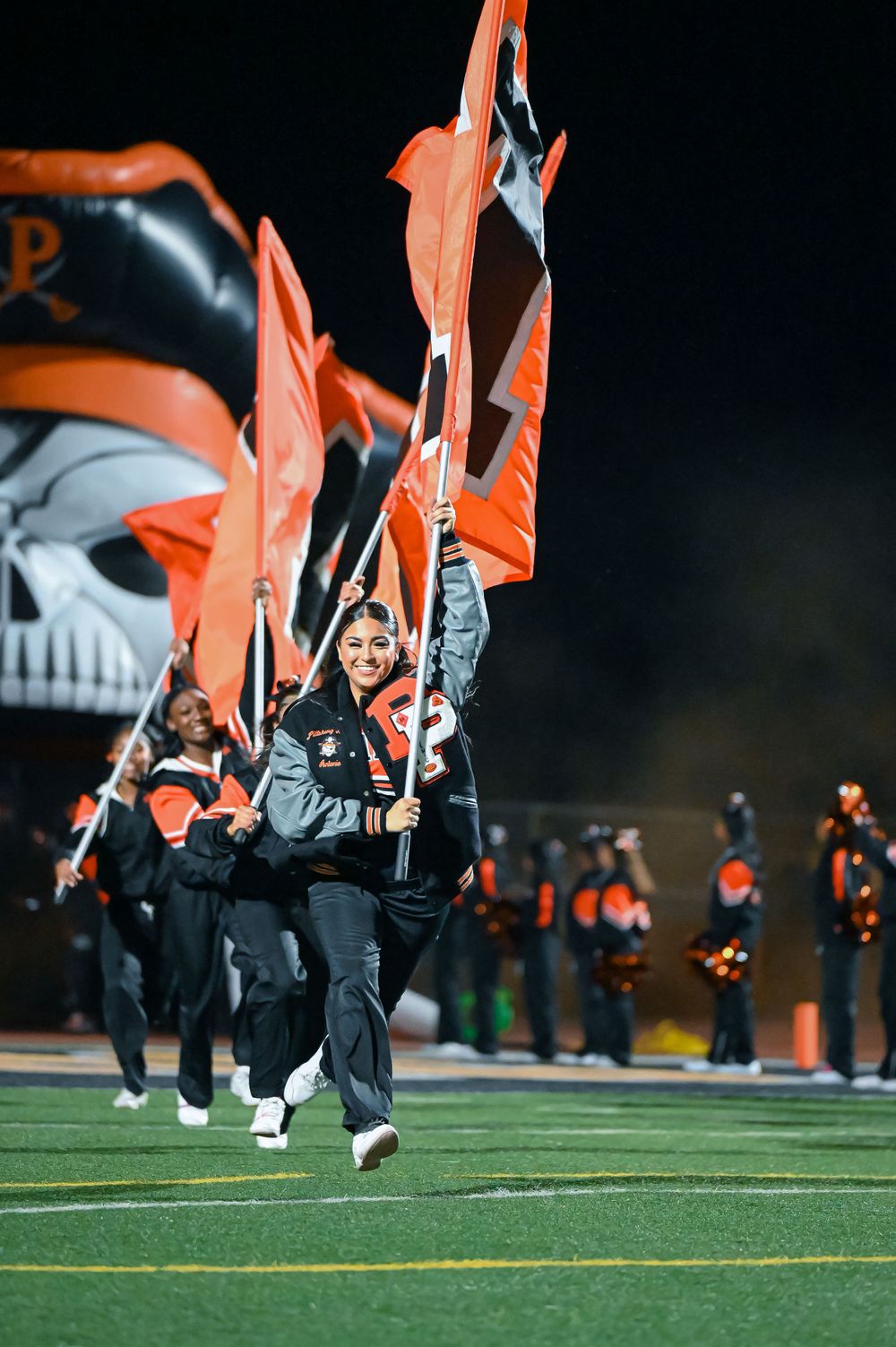 Cheerleaders in black uniforms wave orange flags during a nighttime performance on a football field.