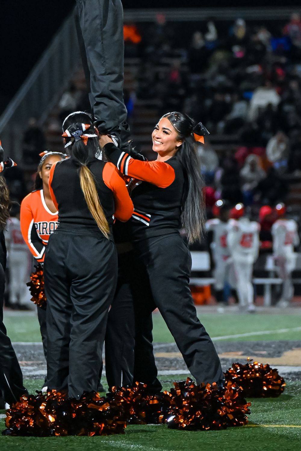 Cheerleaders with orange pom poms perform on the sidelines of a football field at night.