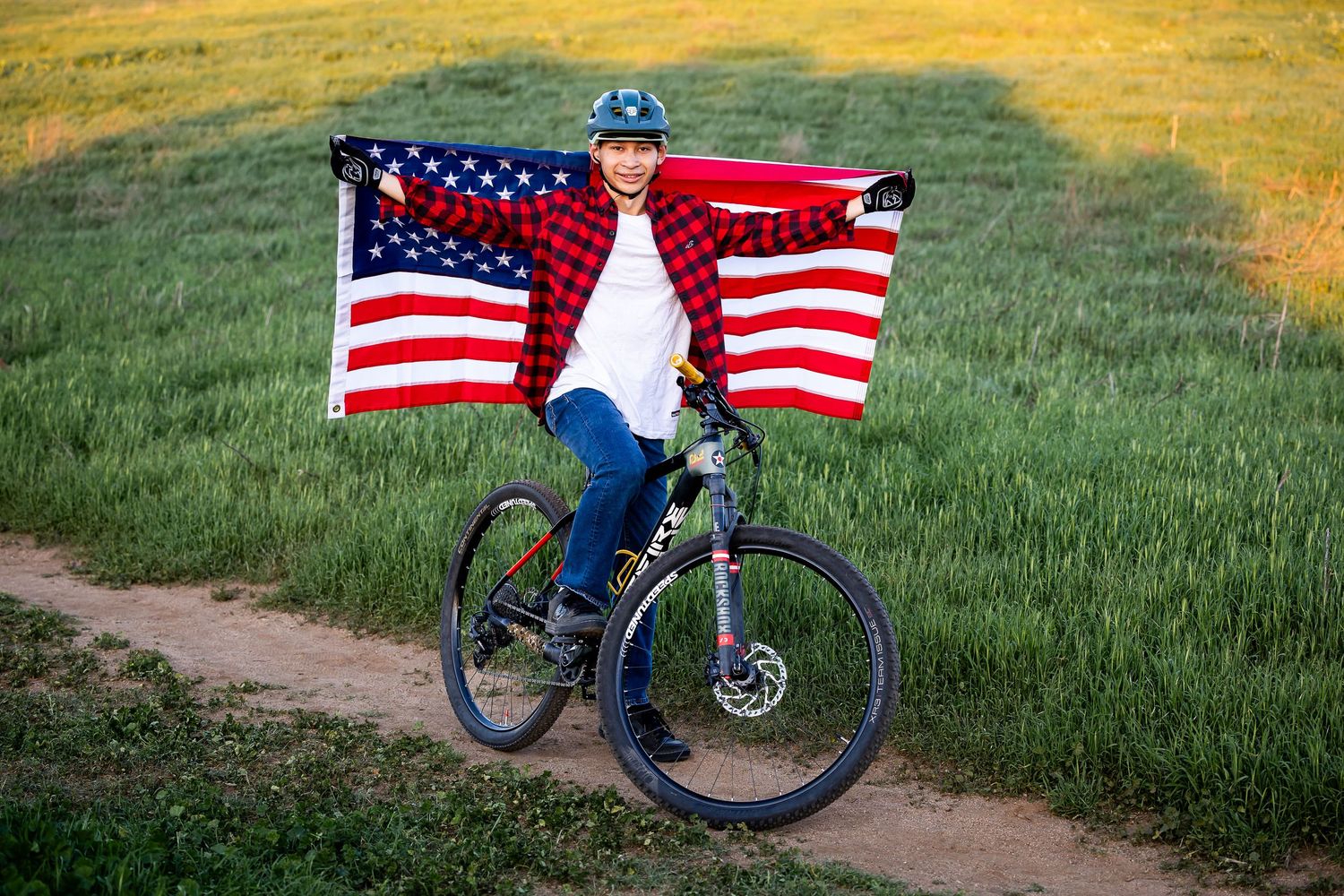 Person with American flag posing with bicycle in golden field at sunset.