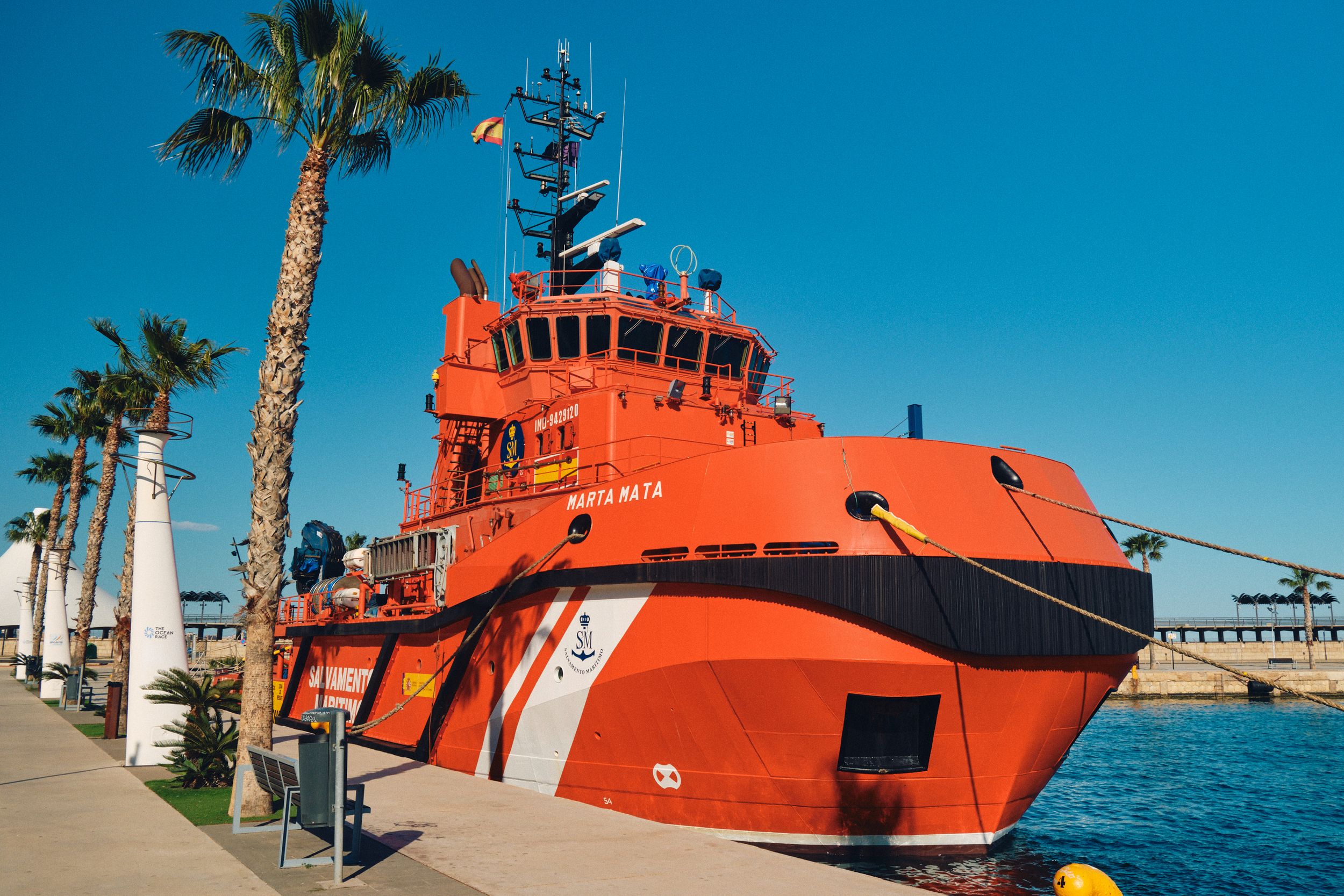 Bright orange rescue ship moored at palm-lined dock under blue sky.