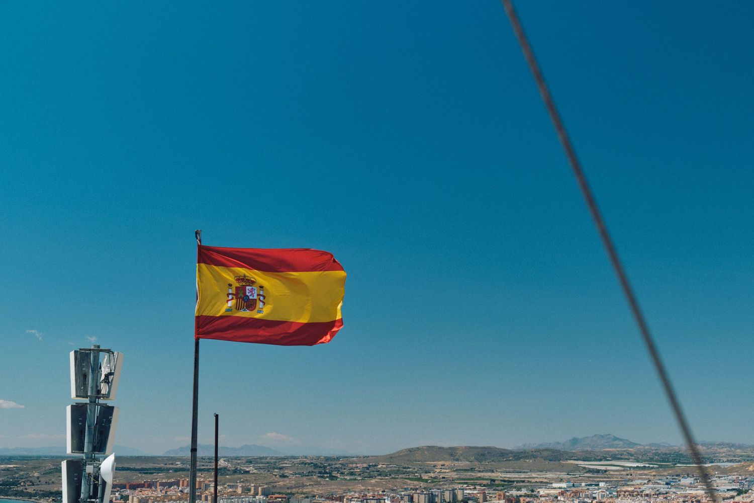 Spanish flag waving against blue sky over Alicante's landscape.
