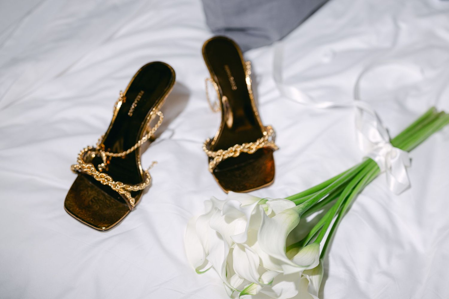 Elegant gold chain-detail sandals next to white flowers on a silky fabric.