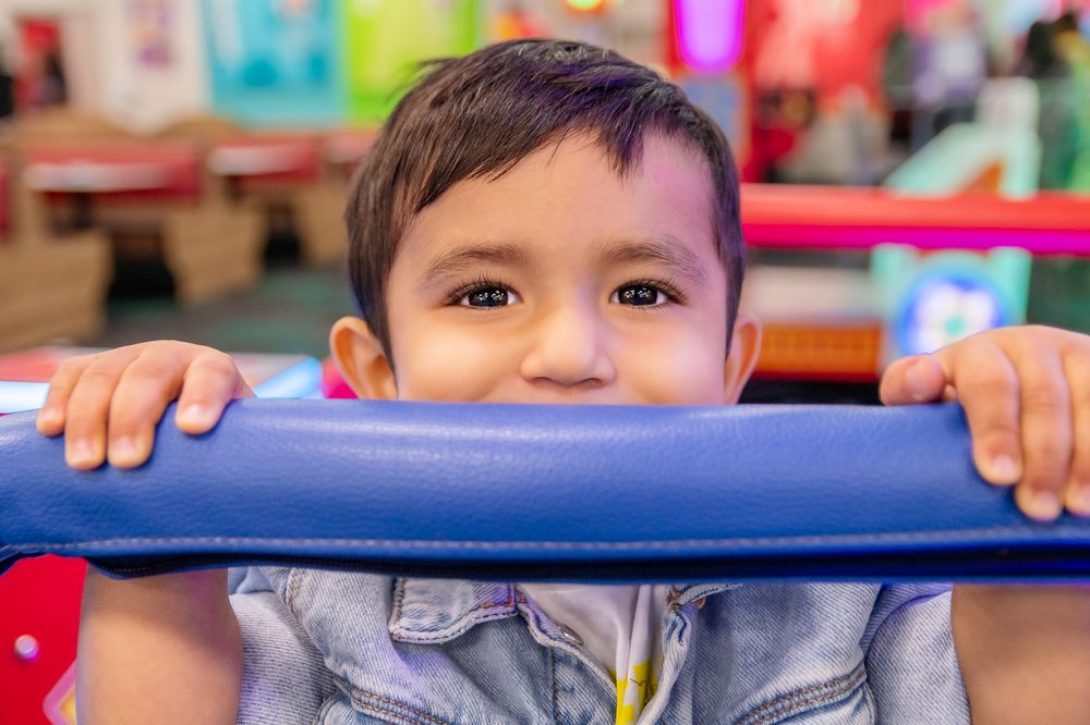 Child playfully peeking over blue cushioned equipment in indoor play area.