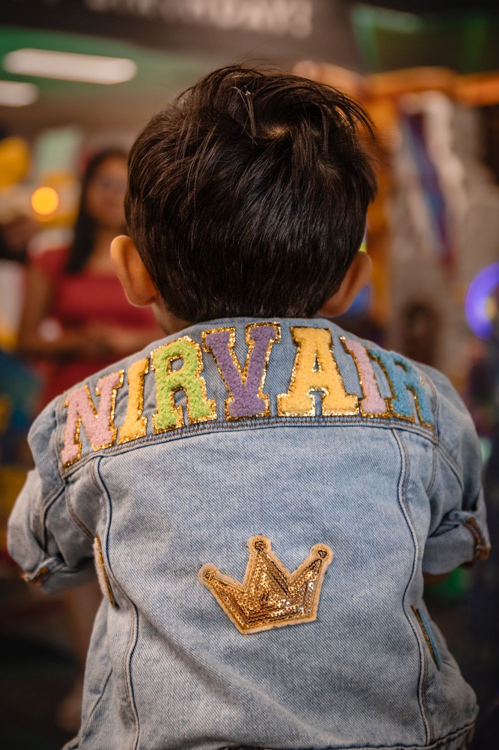 Denim jacket with colorful text and crown embellishment shown from behind.
