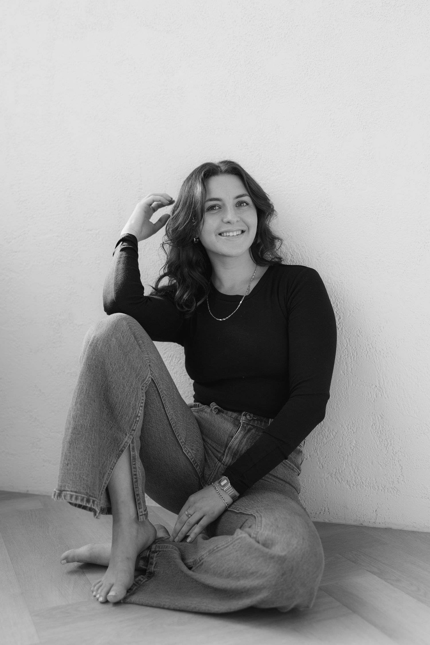 A casual black and white portrait of someone sitting cross-legged against a white wall.