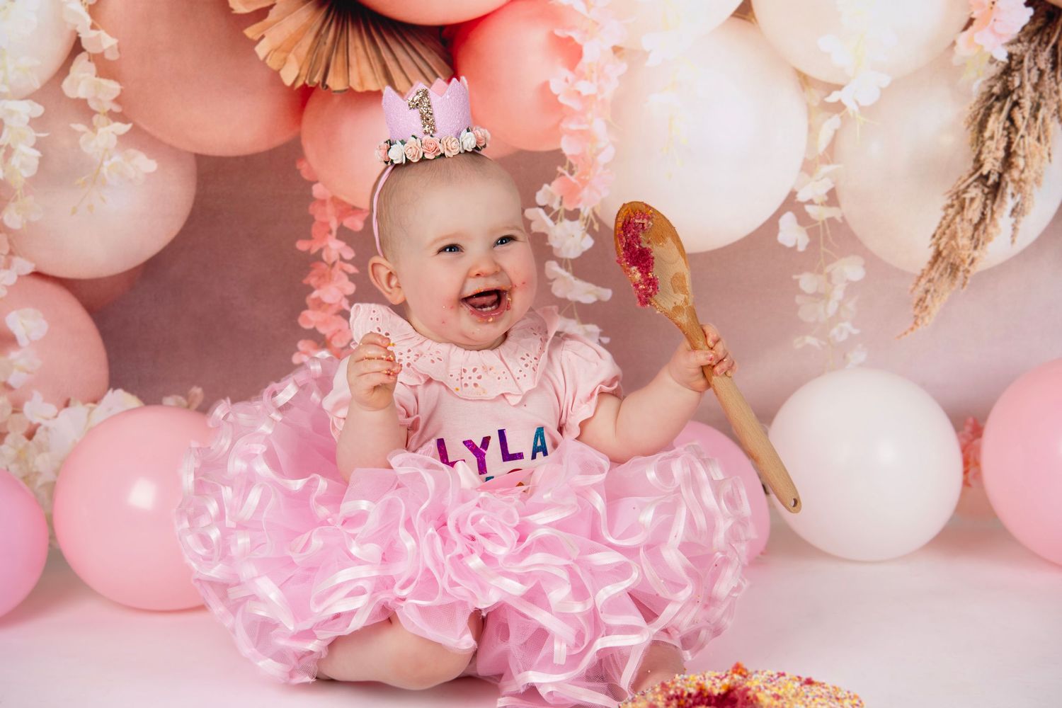 Baby in pink tulle dress with cake smash birthday setup and pink balloon decorations.
