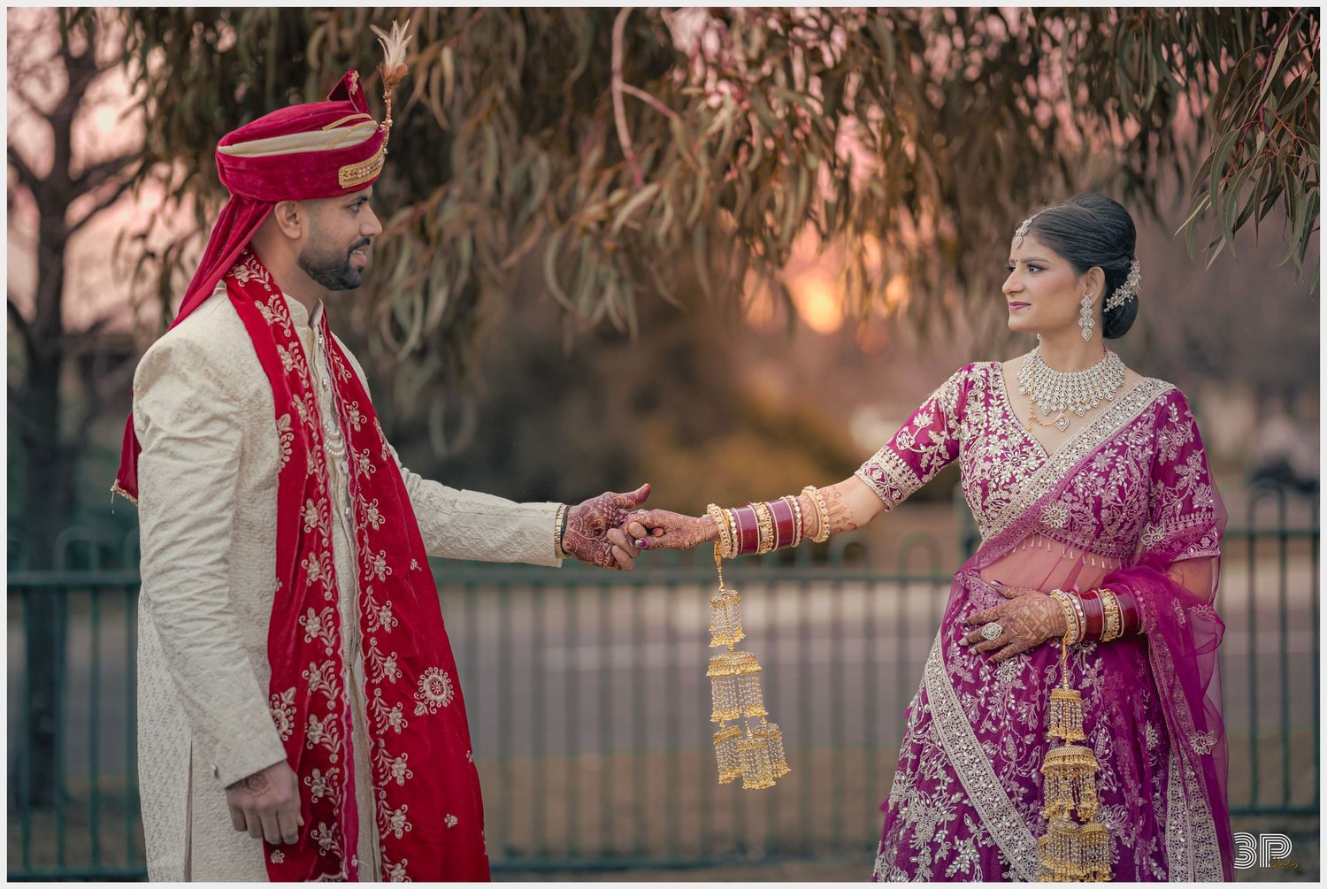 Traditional wedding couple in red and pink attire pose together at sunset under willow trees.