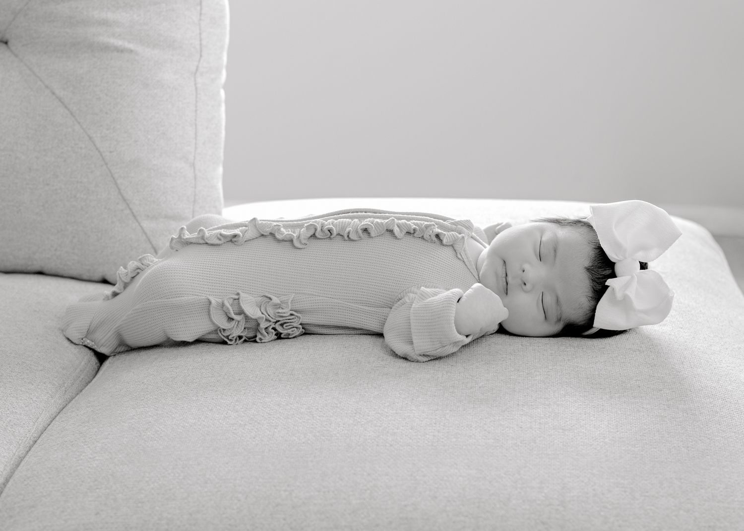 Black and white image of a sleeping baby in ruffled clothing lying peacefully on a gray couch.