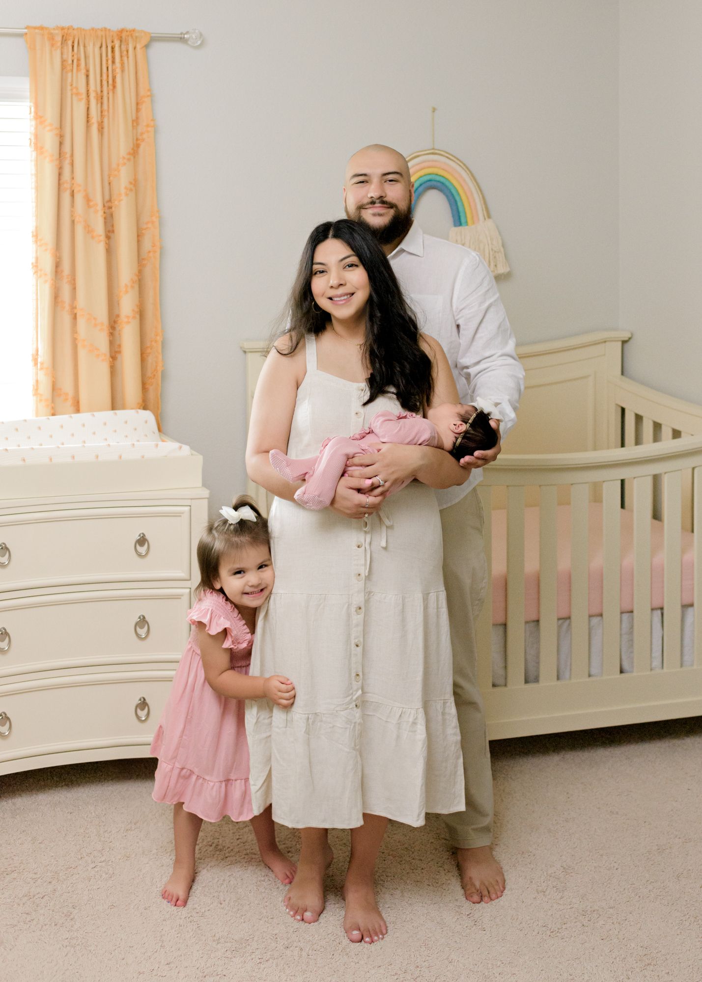 Family portrait in a nursery with rainbow decor and cream-colored furniture.