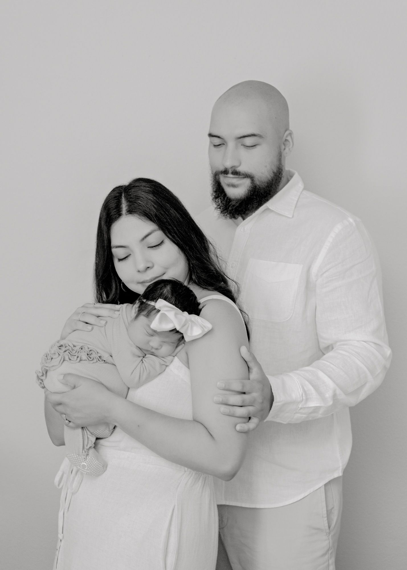 A tender black and white portrait of parents embracing their newborn baby against a light background.