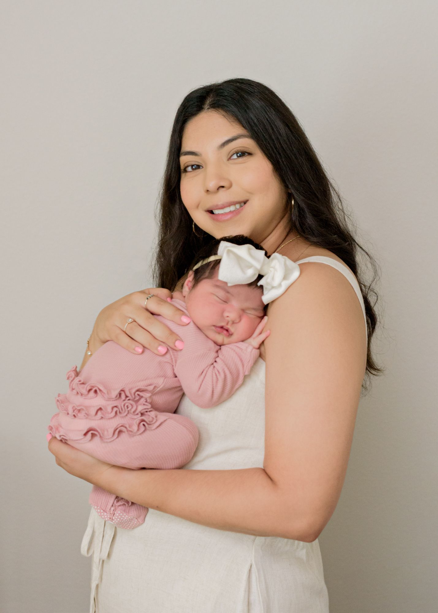 Mother in white dress holding sleeping newborn baby wearing pink ruffled outfit.