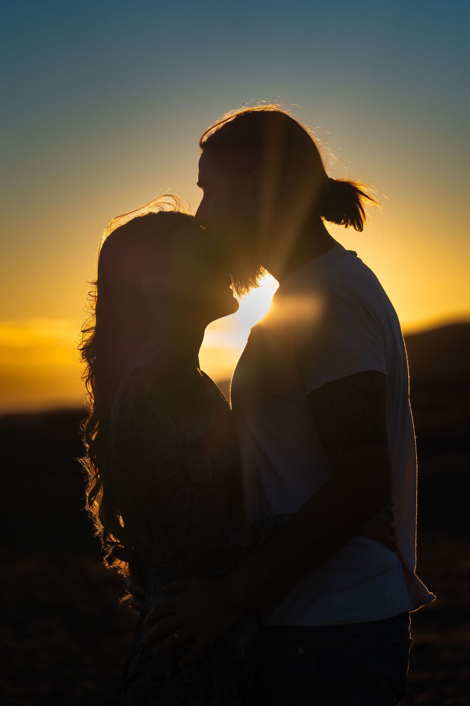 A silhouetted couple shares an intimate moment against a golden sunset sky. Engagement session romantic outdoor photography