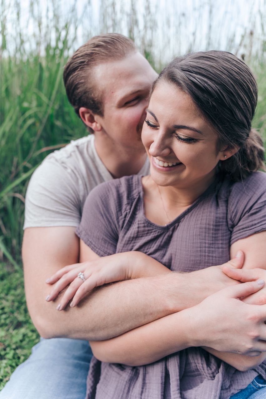 Intimate couple sharing a tender moment outdoors among tall grass with an engagement ring visible. Marion, Ohio