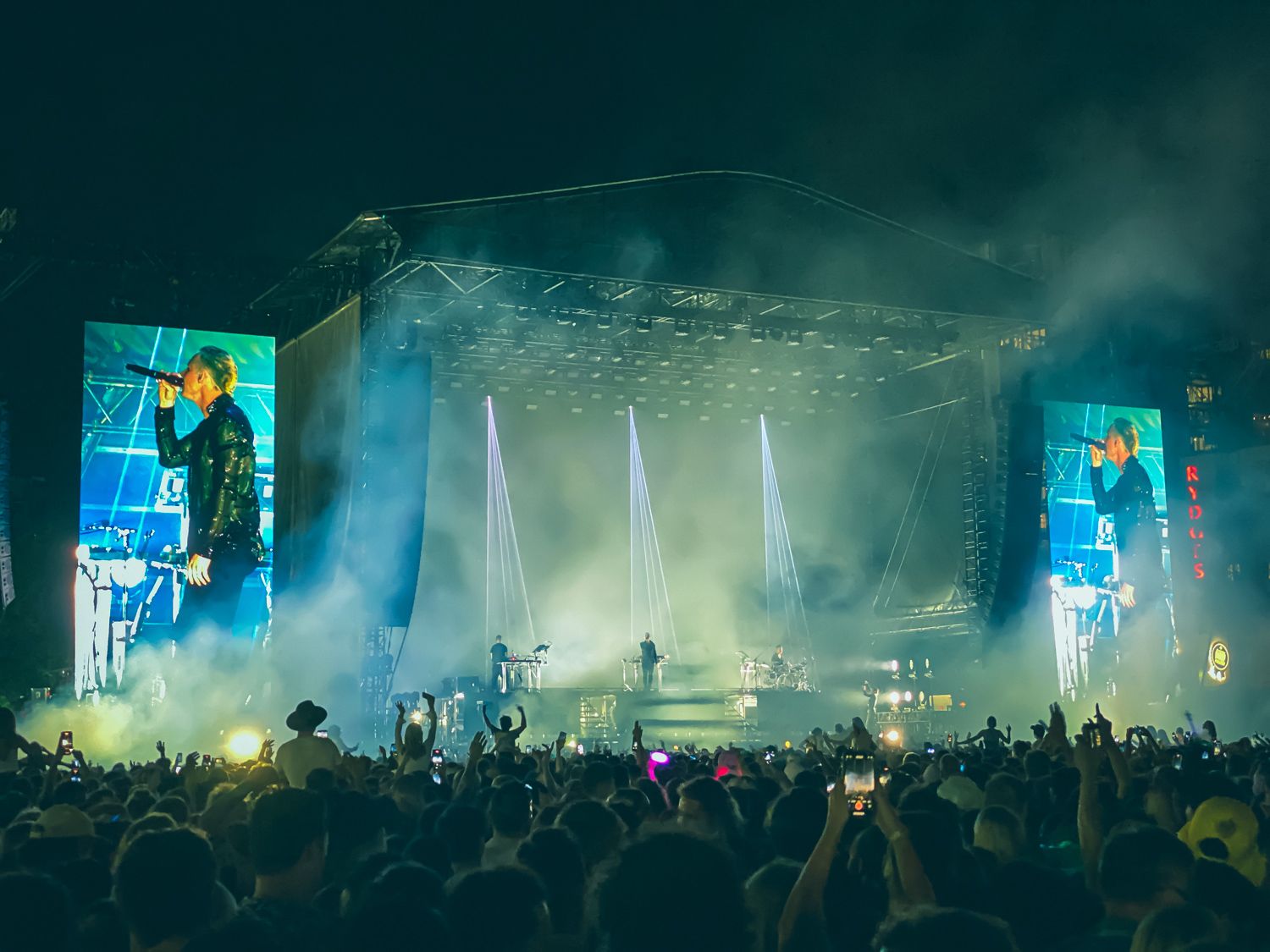 Concert stage with dramatic lighting and fog effects as performer entertains large crowd at night.