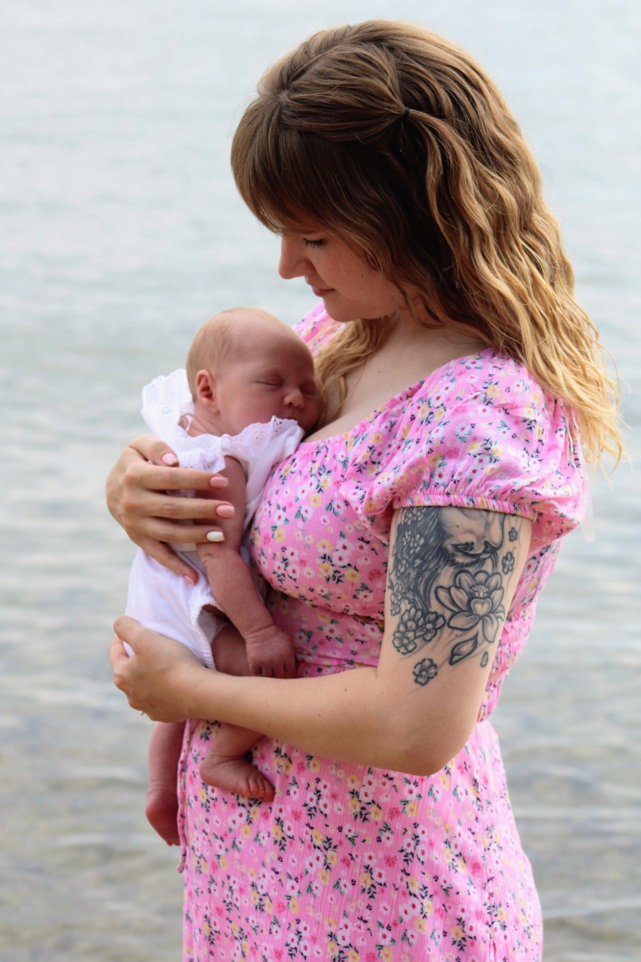 Young mother in pink floral dress with tattoo tenderly holds newborn baby by the ocean shore.