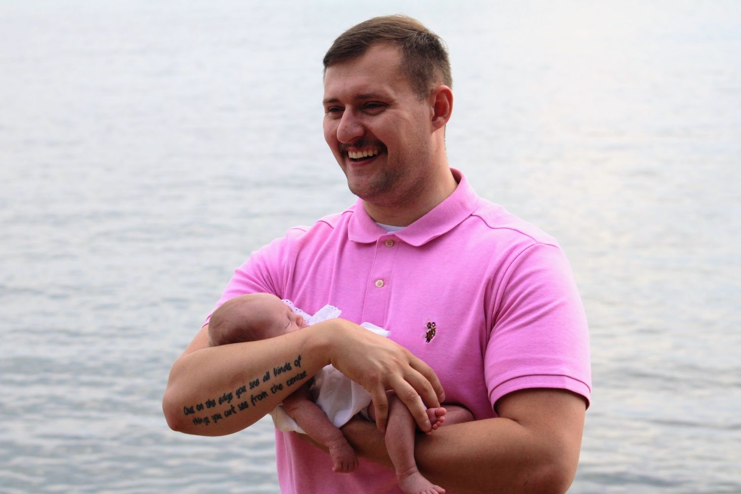 Man in pink polo shirt holding a newborn baby near the ocean shore, smiling outdoors.