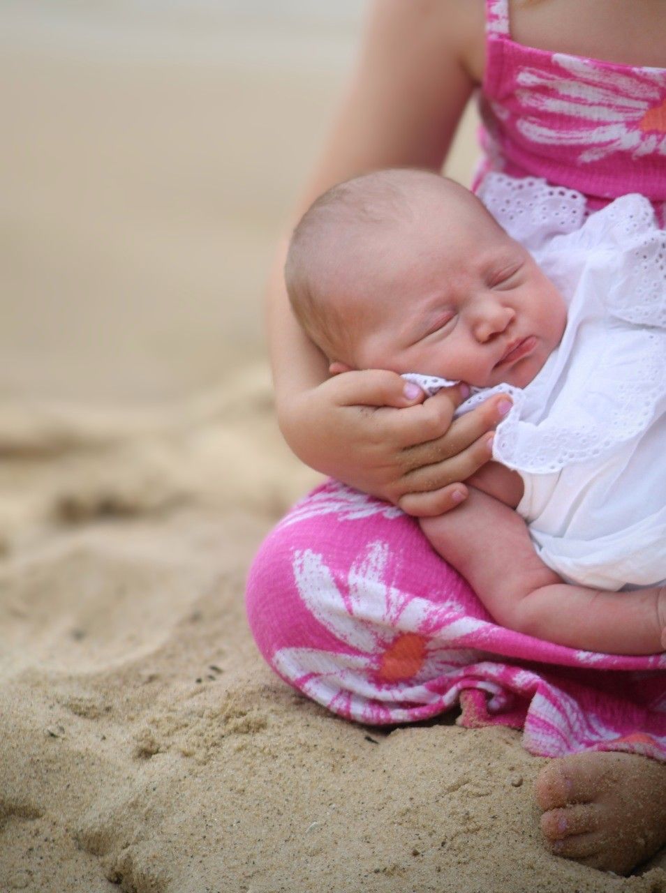 A sleeping newborn baby in white lace outfit cradled by mother in pink floral dress on sandy beach.