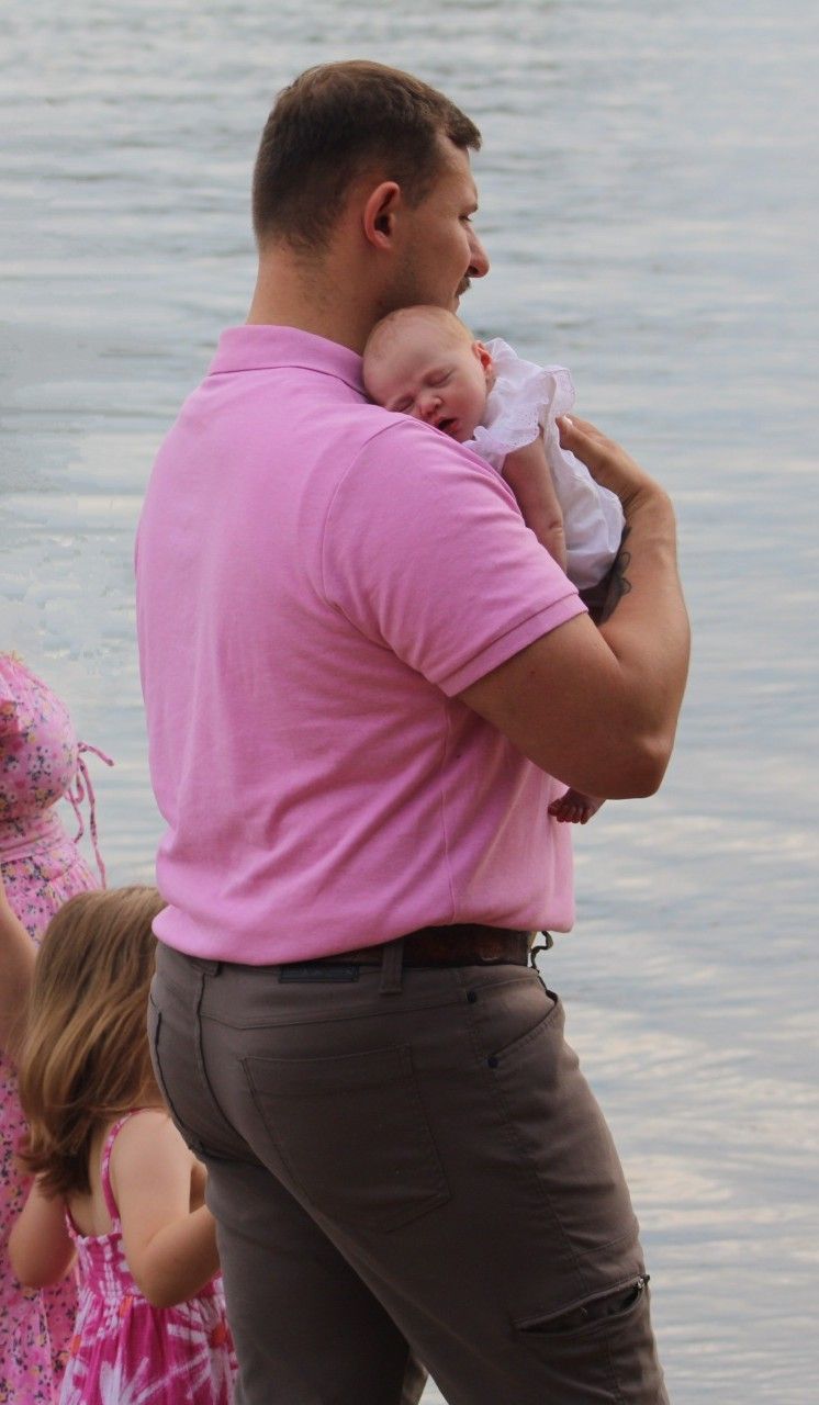 Man in pink polo shirt holding a newborn baby near a waterfront with a young girl standing nearby.