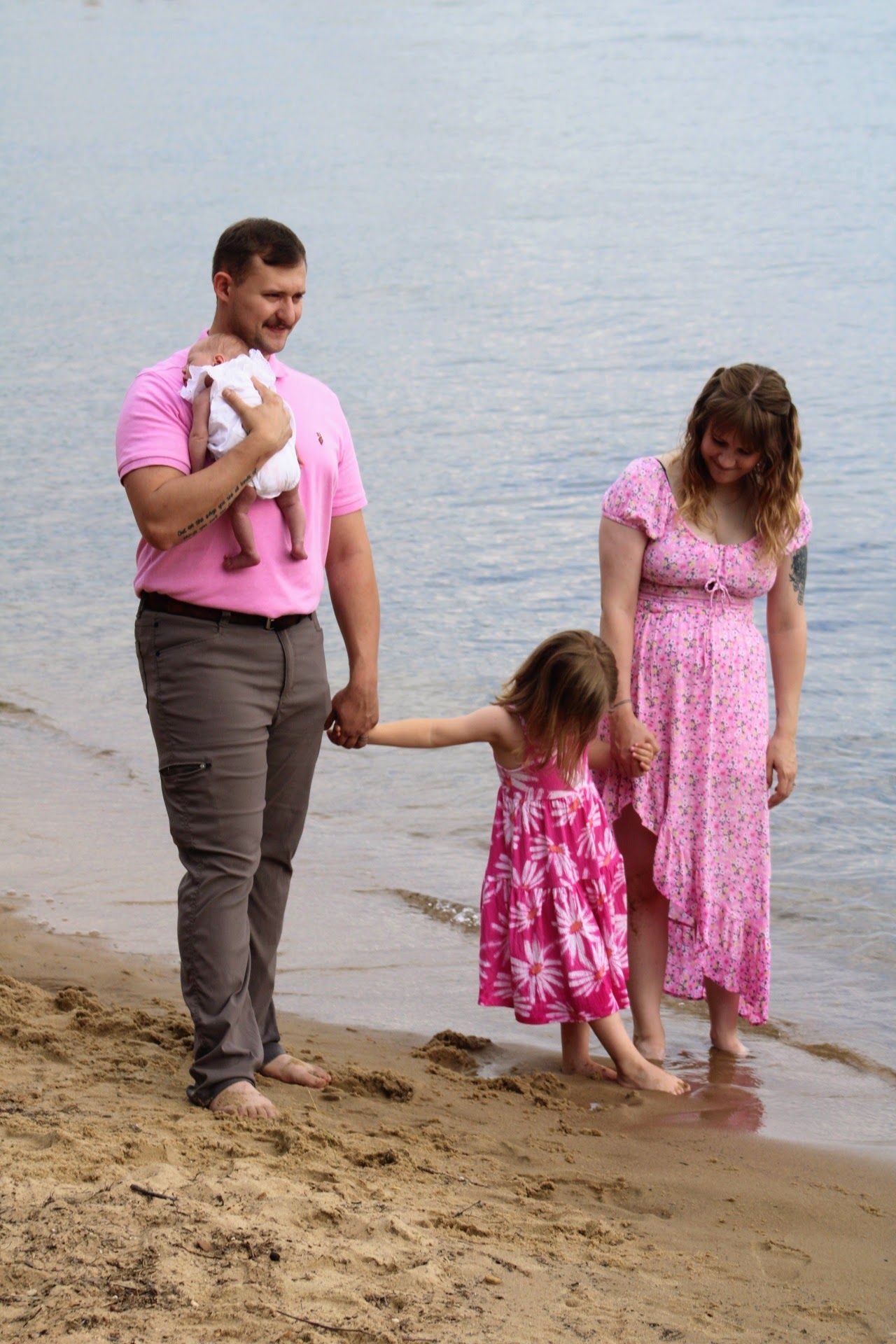 Family in pink outfits on sandy beach, father holding baby, mother and young daughter paddling in water.