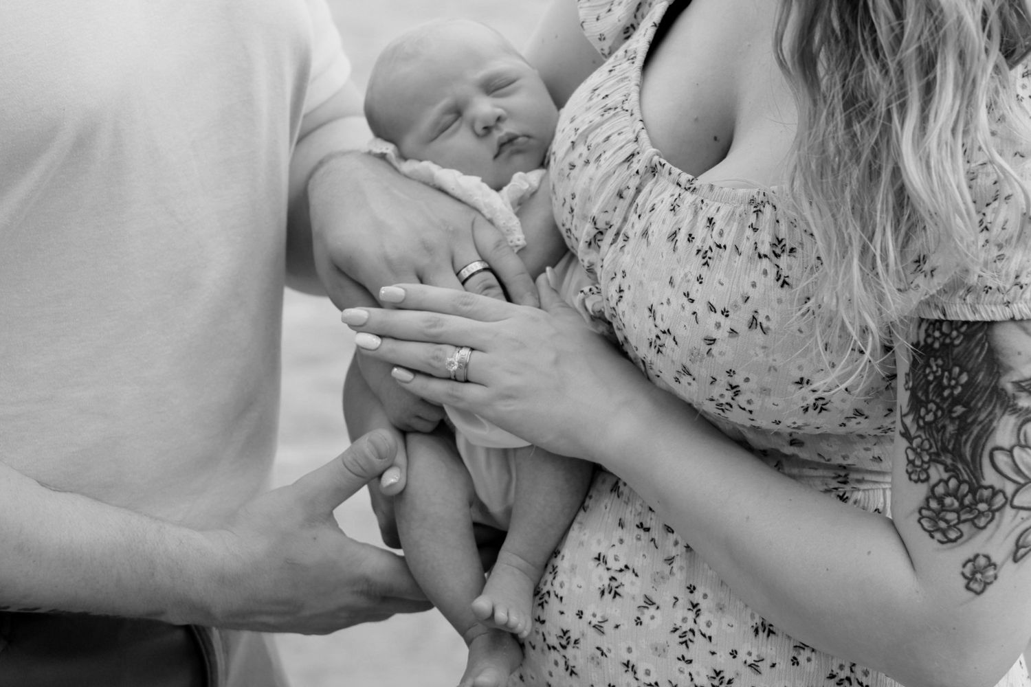 Black and white photo of parents cradling their newborn baby, mother wearing floral dress with tattoo visible.