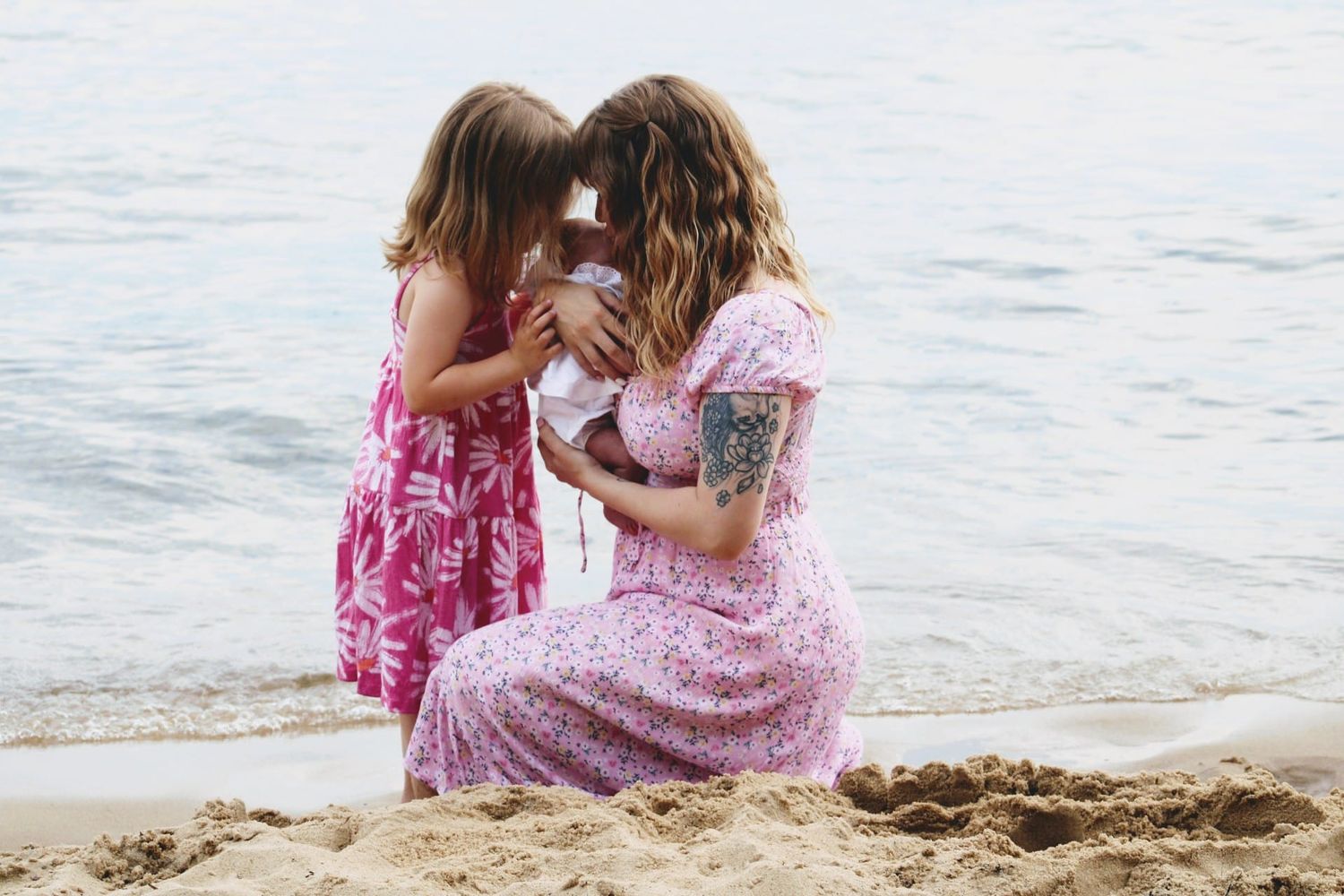 A mother and young daughter in pink dresses share a tender moment with baby on a sandy beach by calm waters.