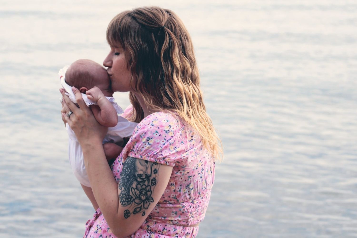 Tattooed mother in pink floral dress kissing newborn baby by the ocean shore.