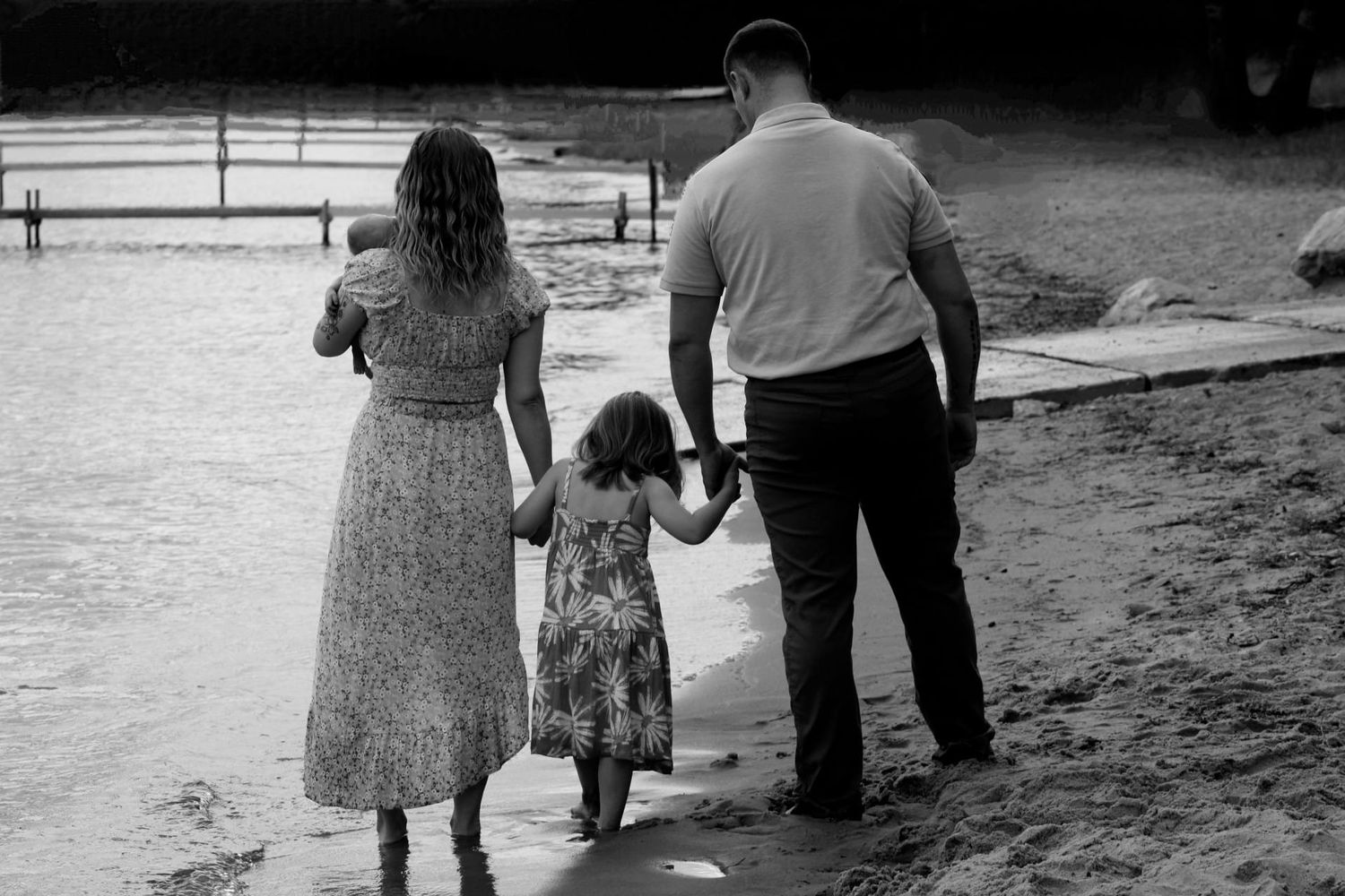 Black and white photo of a family holding hands by a lake shore, parents and young child standing together.