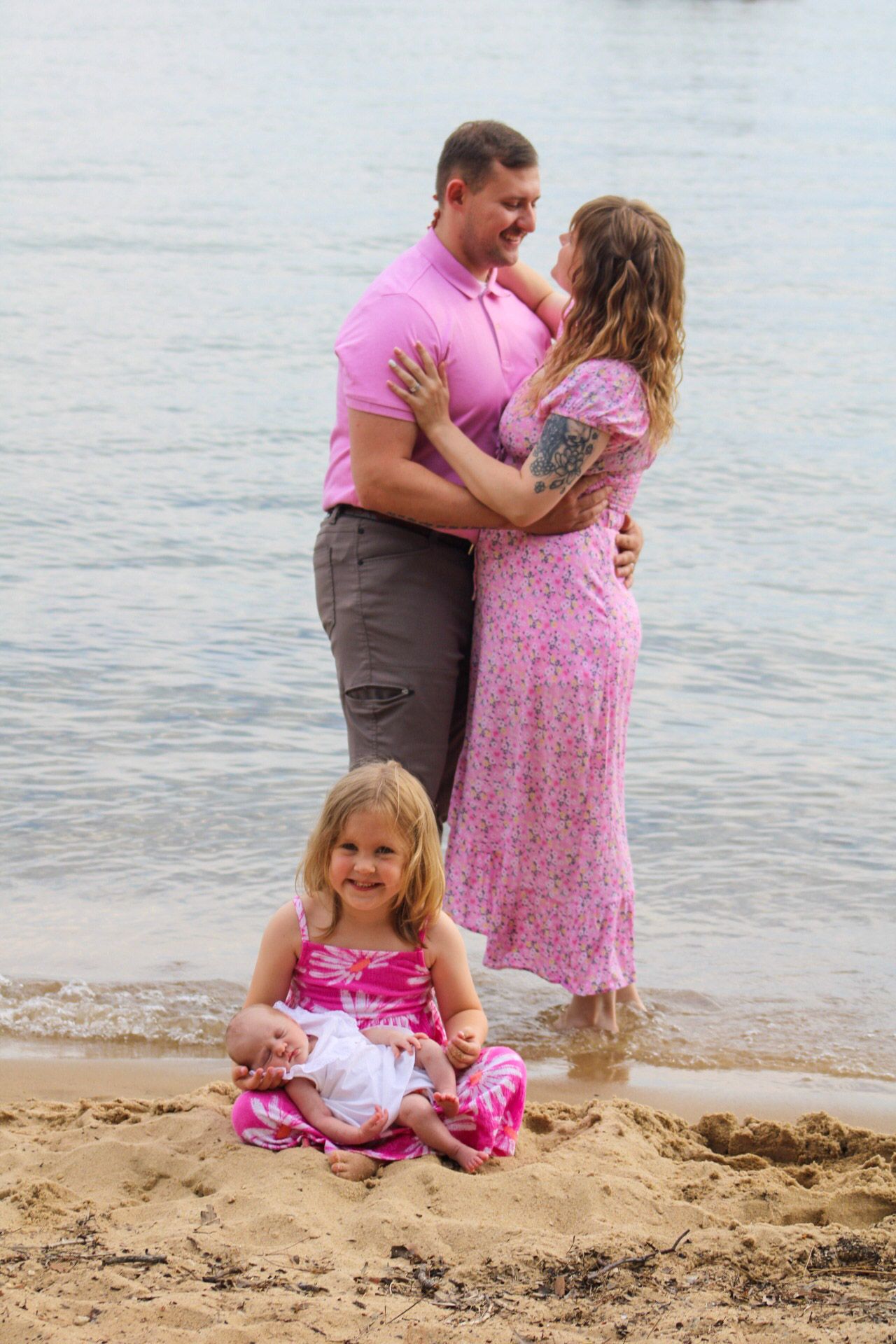 Family beach photo with couple embracing near water as young girl in pink dress sits on sandy shore.