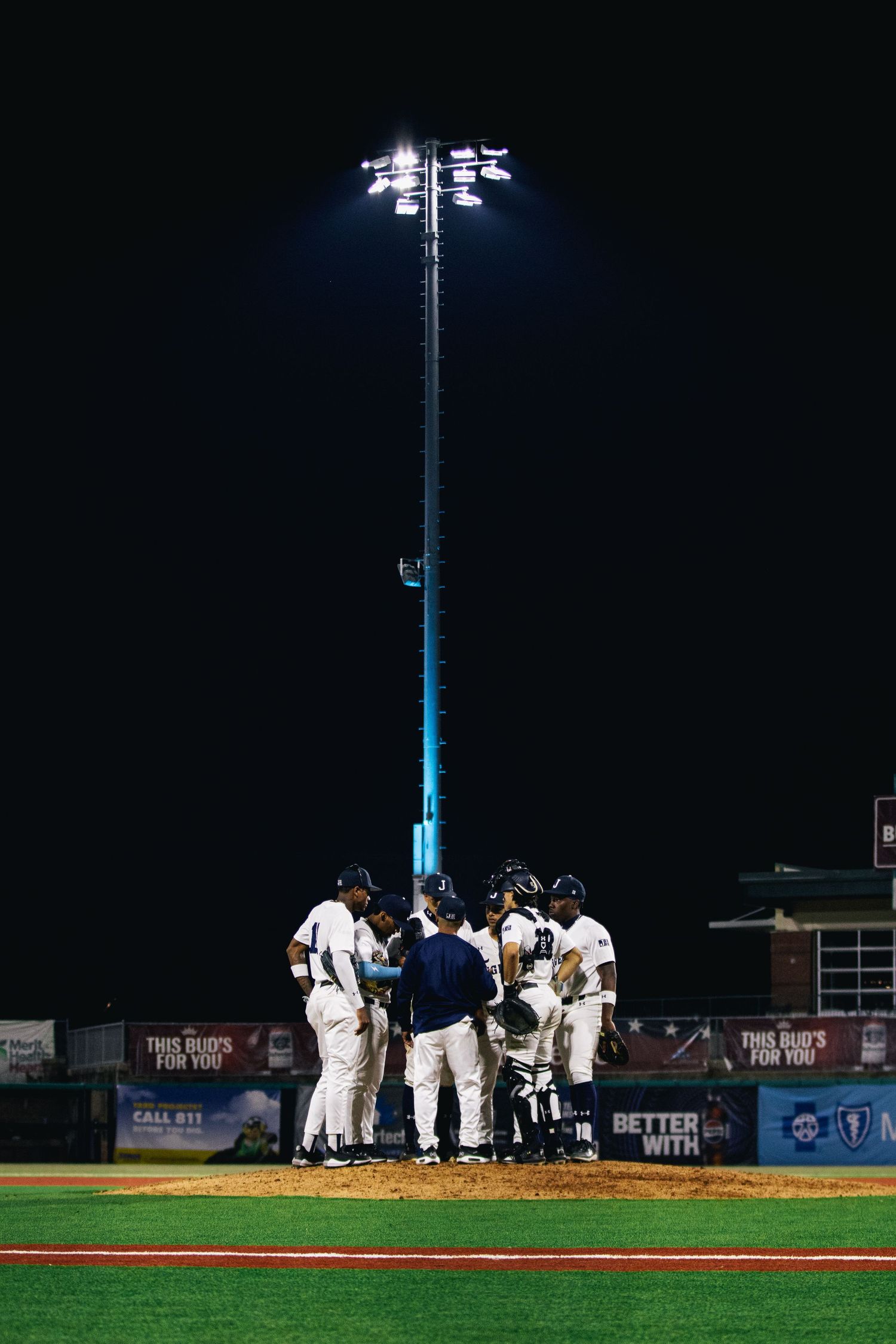 Baseball team huddles on the pitcher's mound under bright stadium lights at night.