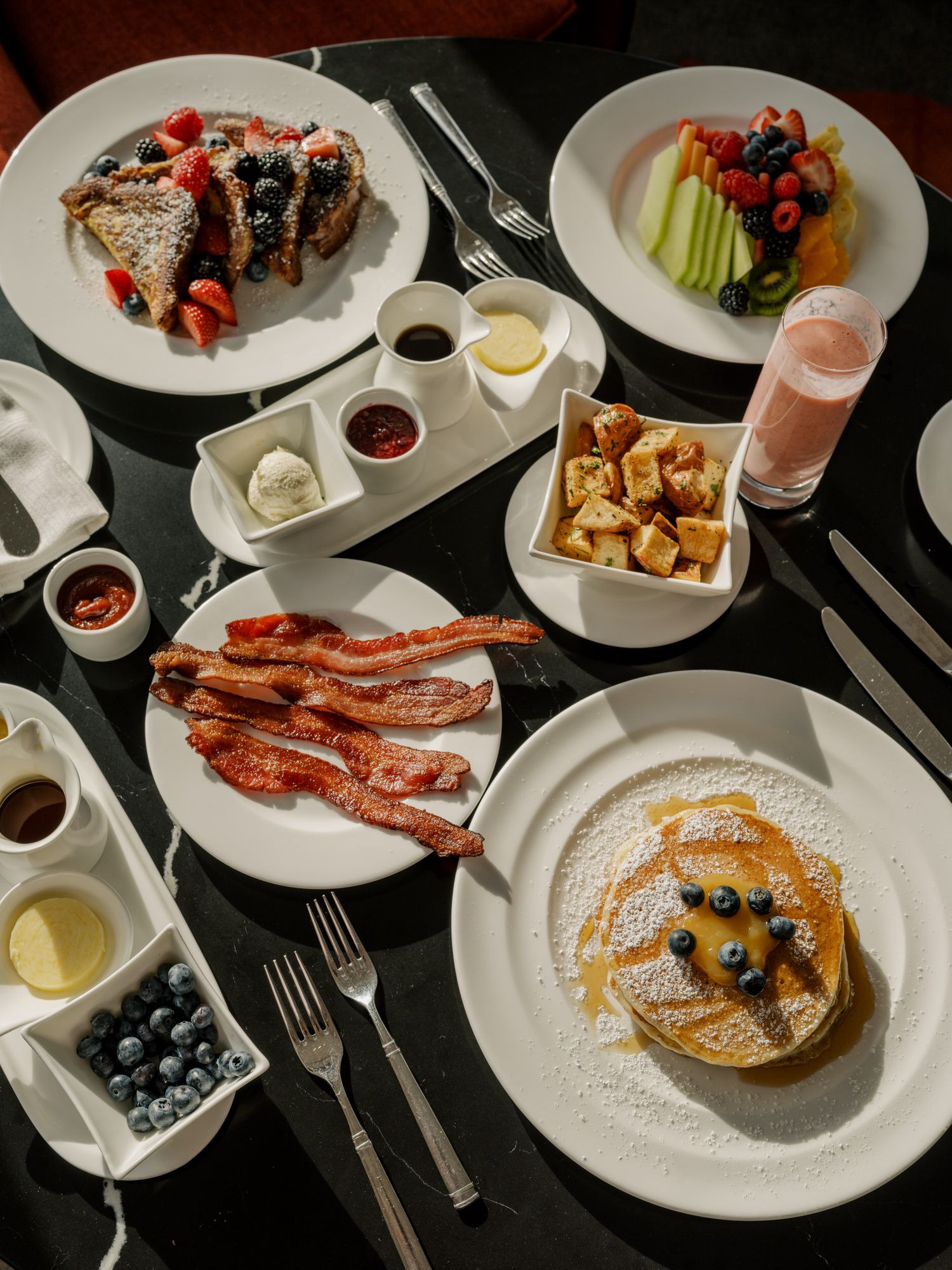 Brunch spread featuring fluffy pancakes with fresh berries, crispy bacon strips, and assorted breakfast sides on white plates.