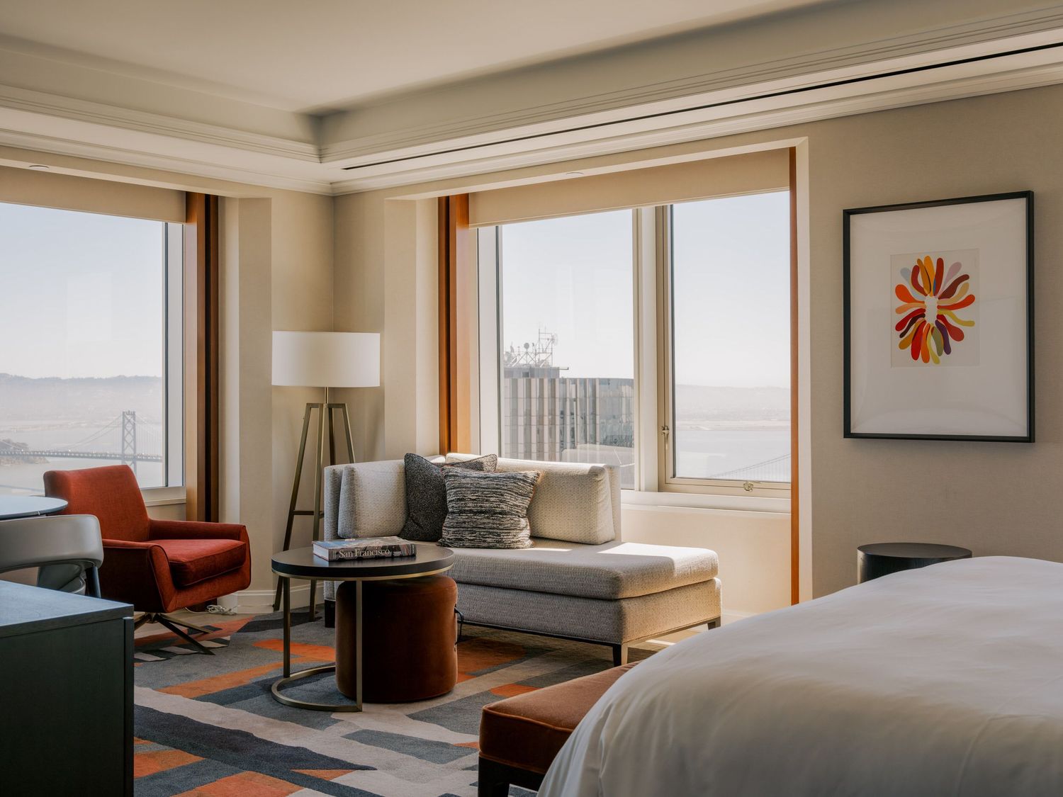Modern hotel room interior with large windows, chaise lounge, orange accent chair and contemporary wall art.
