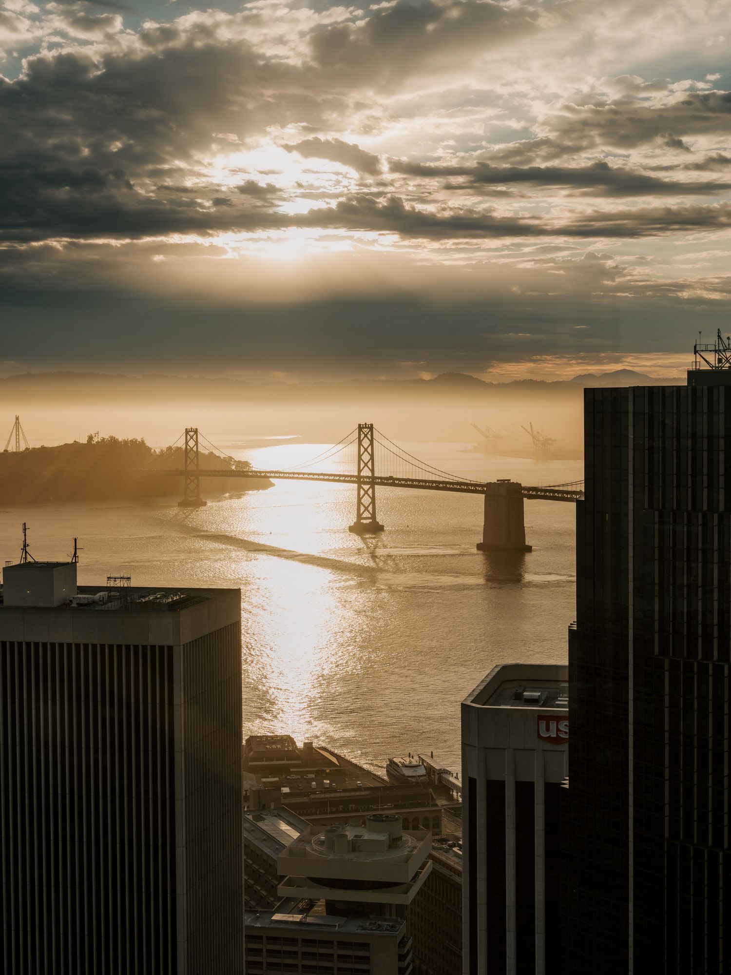 Aerial view of the Bay Bridge in San Francisco at sunset with dramatic clouds and golden light reflecting on the water.