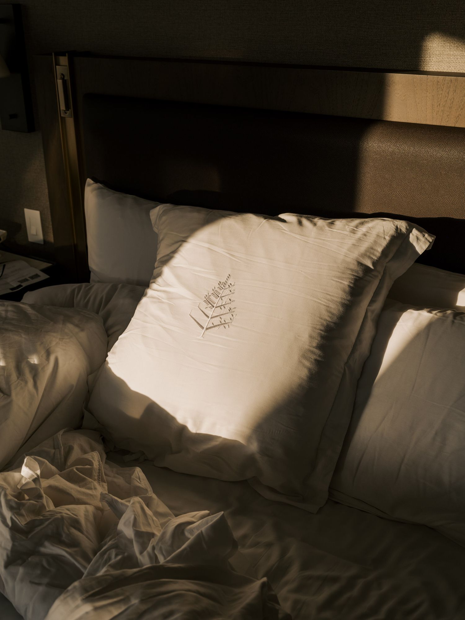 White decorative pillow with embroidered Four Seasons leaf logo design on a bed in moody lighting.