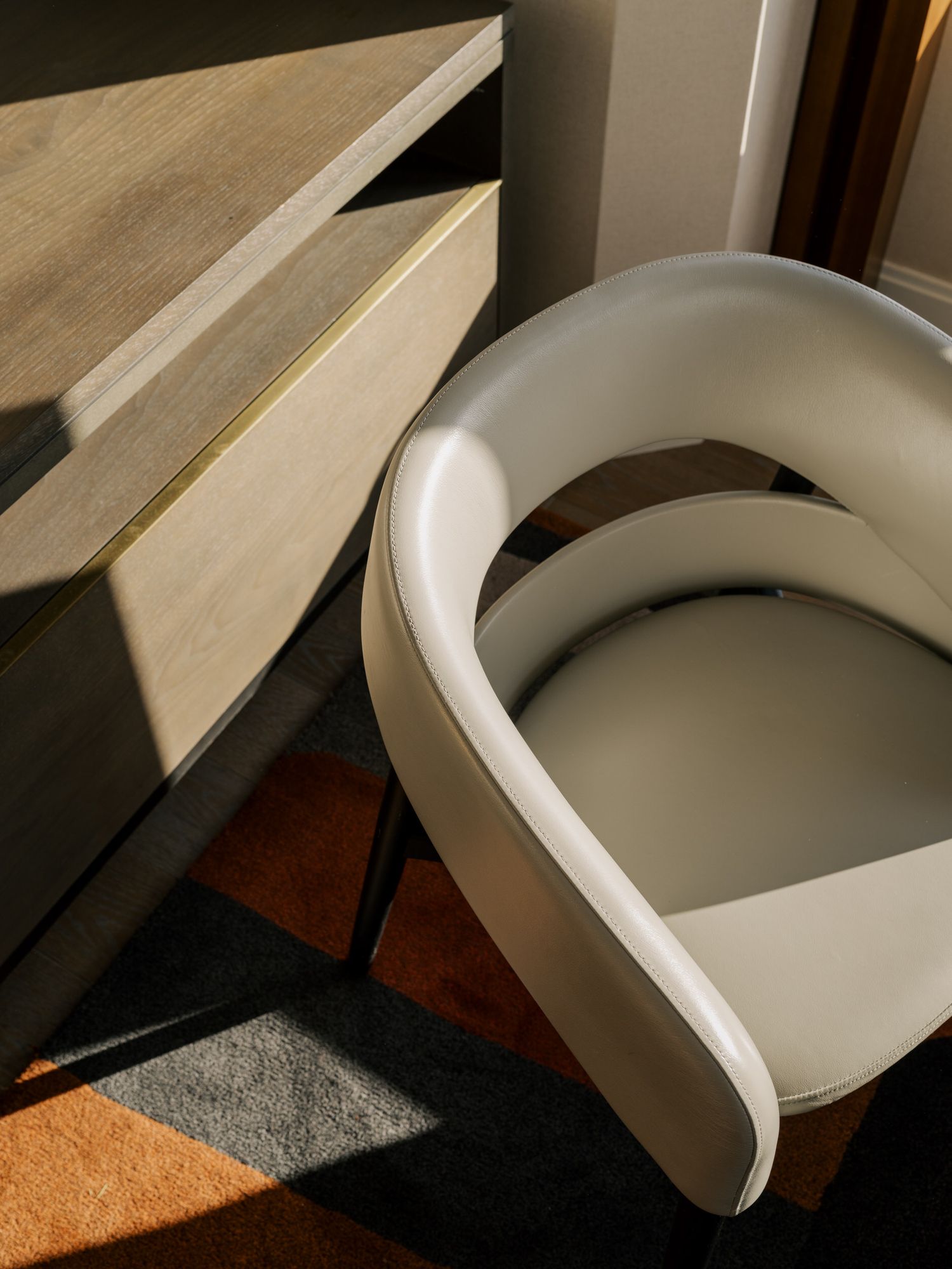 A modern white curved accent chair sits beside wooden furniture in a sunlit corner with dramatic shadows.