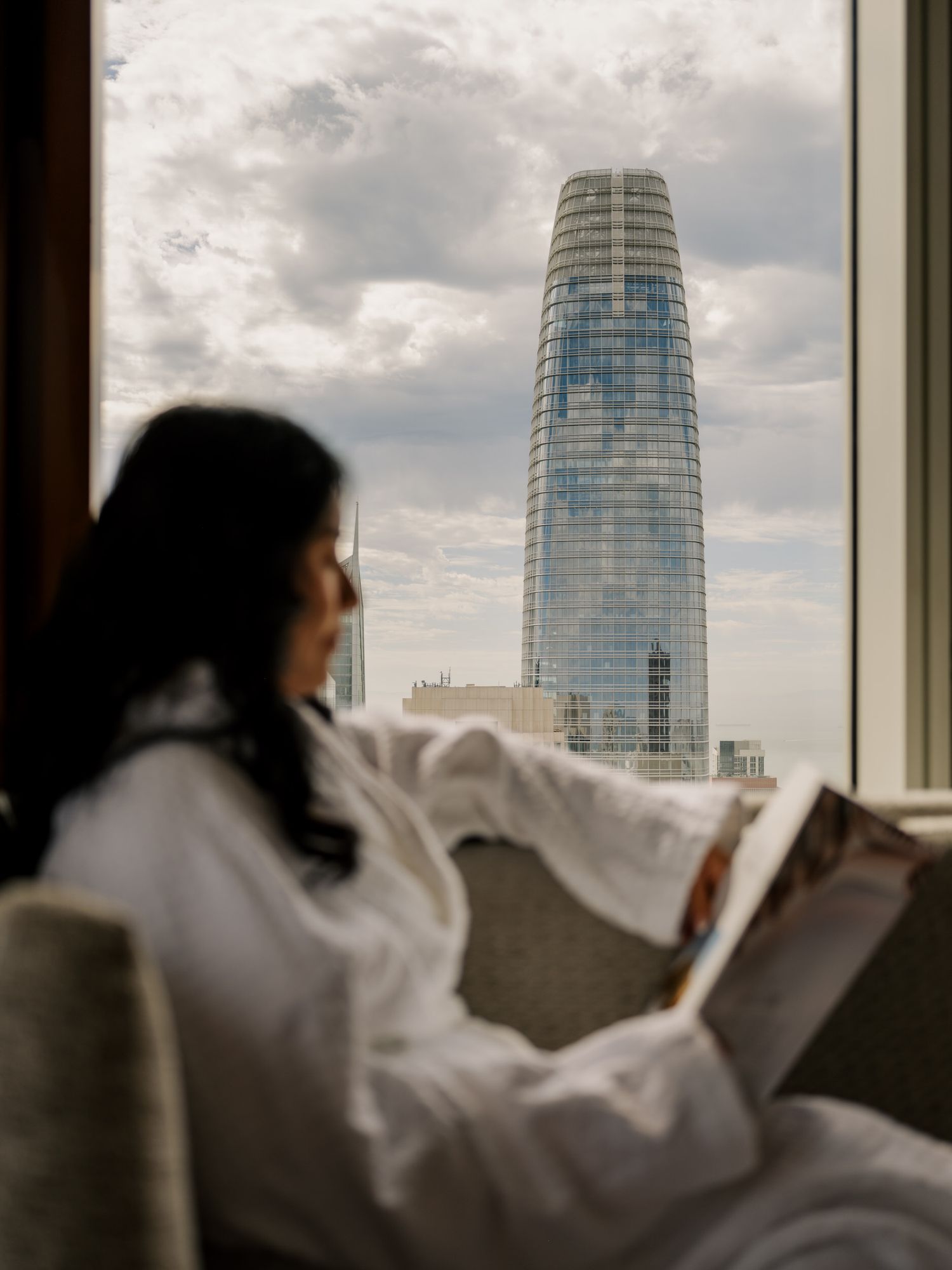 Woman relaxes in robe while looking out of her San Francisco cityview hotel room at the Four Seasons Embarcadero property.