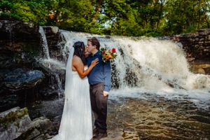 A romantic wedding kiss captured in front of a rushing waterfall on a sunny day.