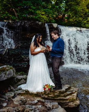 A ceremony takes place on rocky terrain with a beautiful waterfall backdrop in a forest setting.